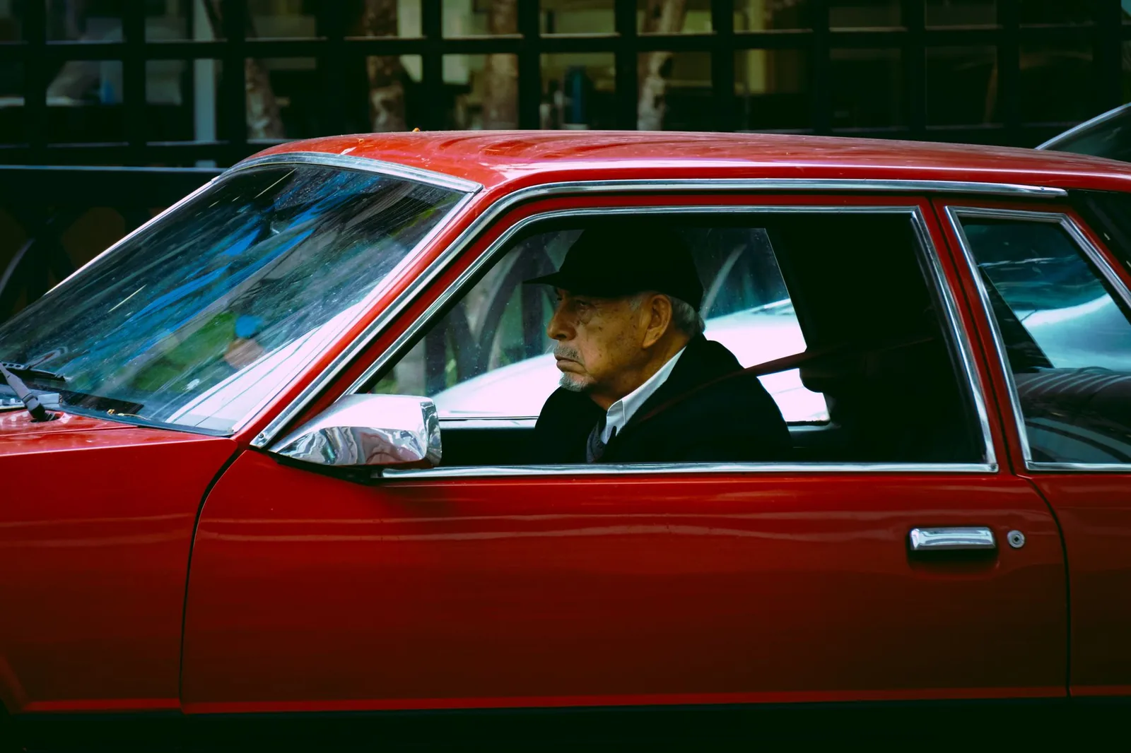 An older man in a hat driving a vintage red car, seen in profile through the driver's side window.