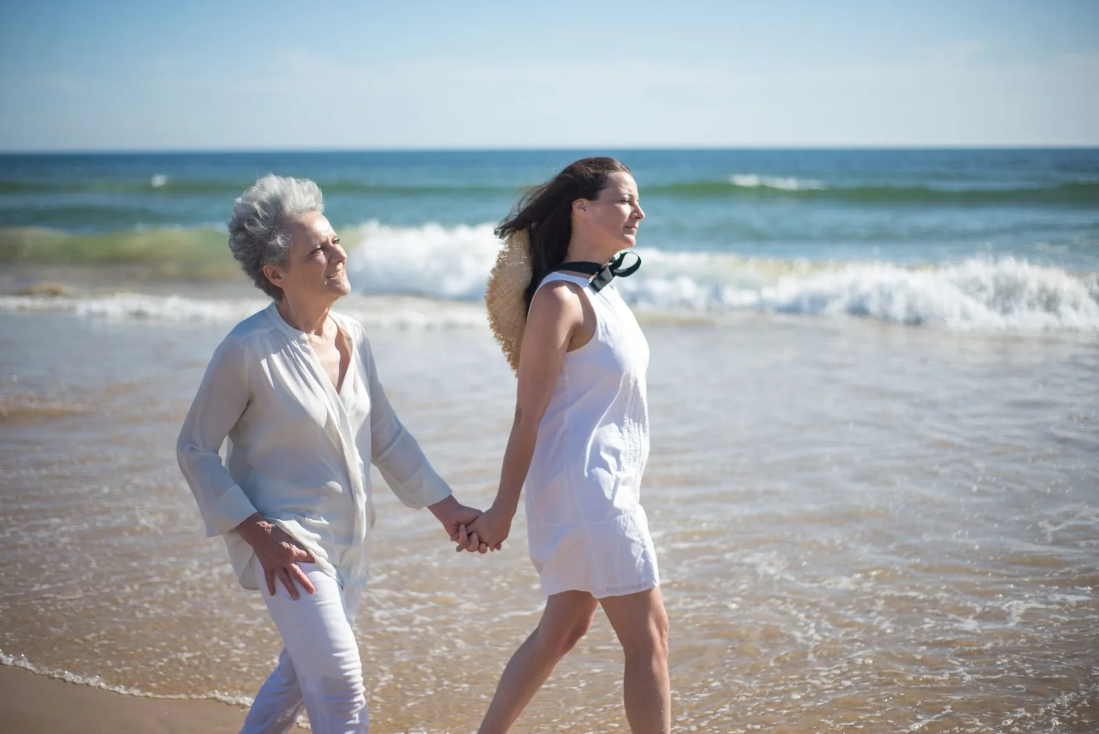 A senior woman and her daughter holding hands while walking along a sunny beach.