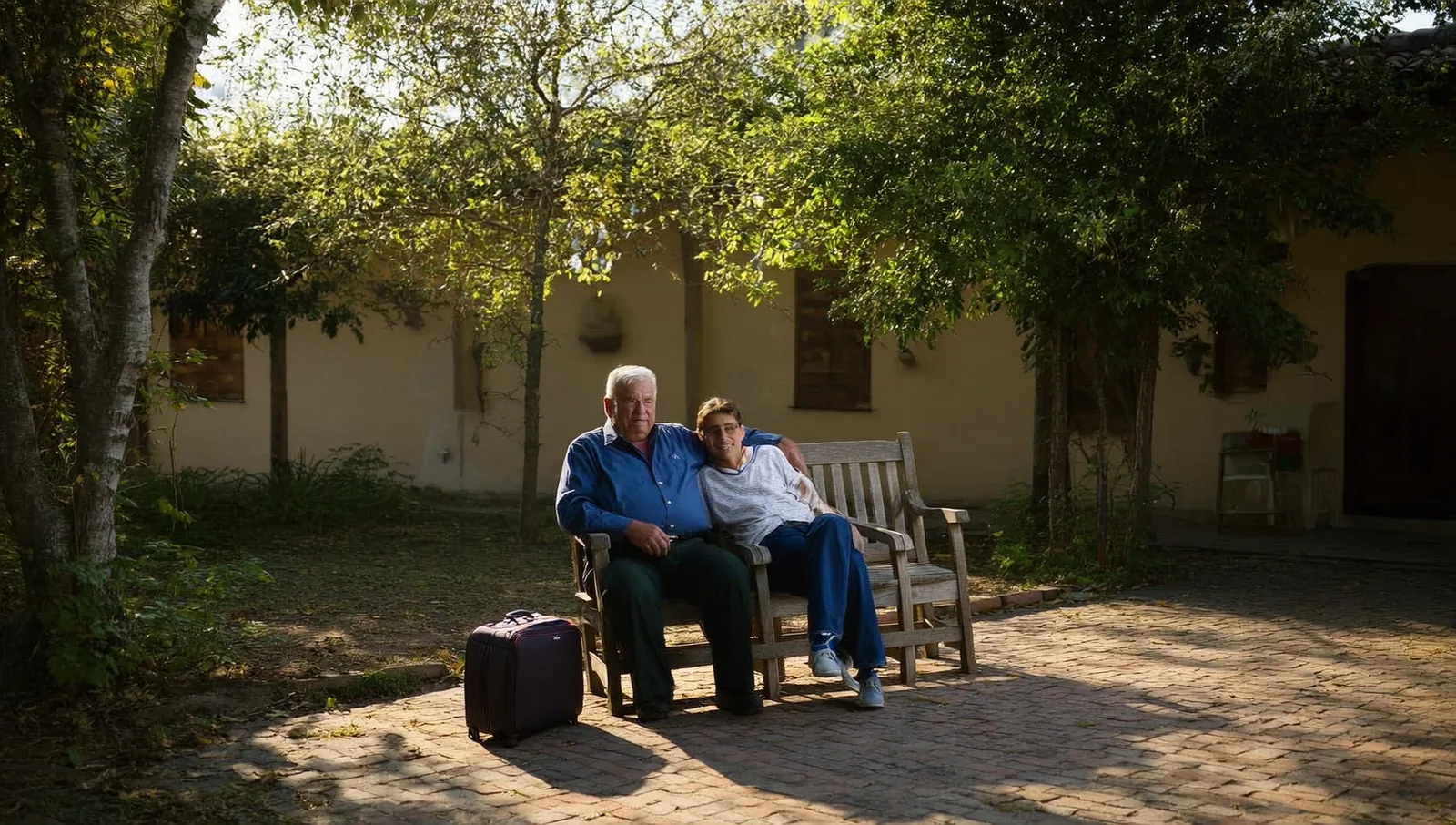An older man and a grown family member sit close together on a wooden bench in a sunlit garden courtyard, a small suitcase on the ground beside them.