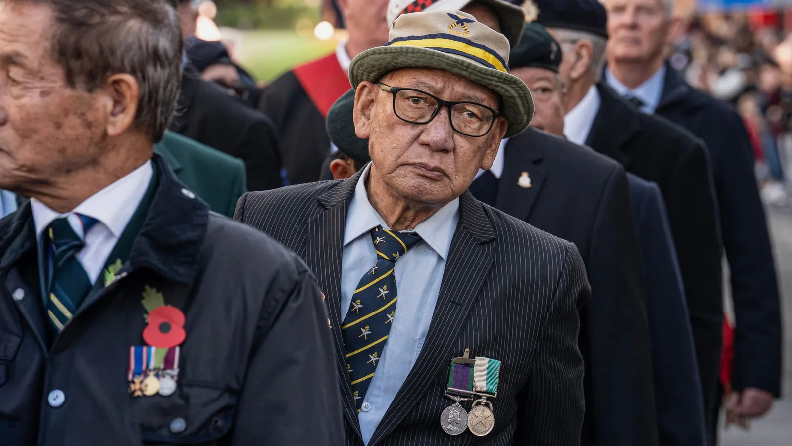 Elderly veterans in suits and service medals marching together in a Remembrance Day parade.
